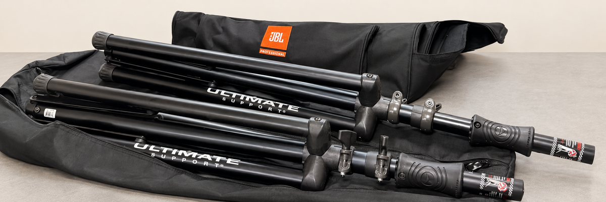 Two actual folded Ultimate Support speaker stands with bags on a clean staged neutral background.